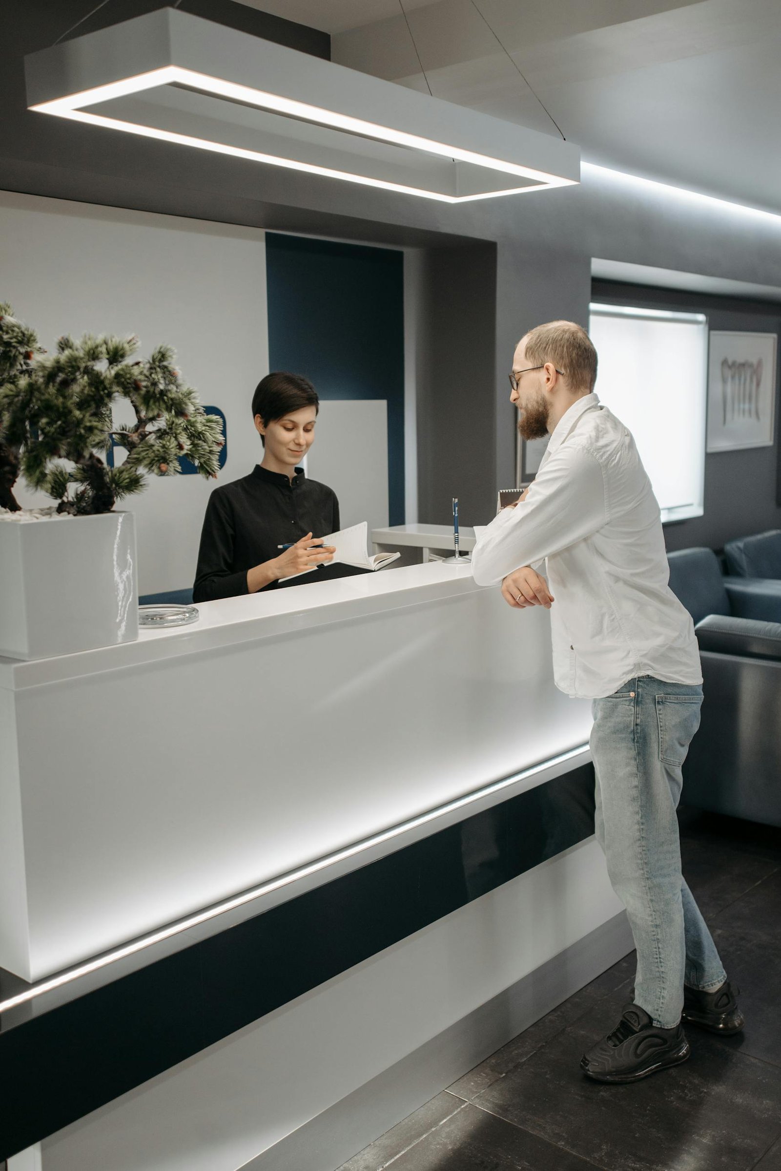 A man and woman at a sleek office reception desk, discussing paperwork in a professional setting.